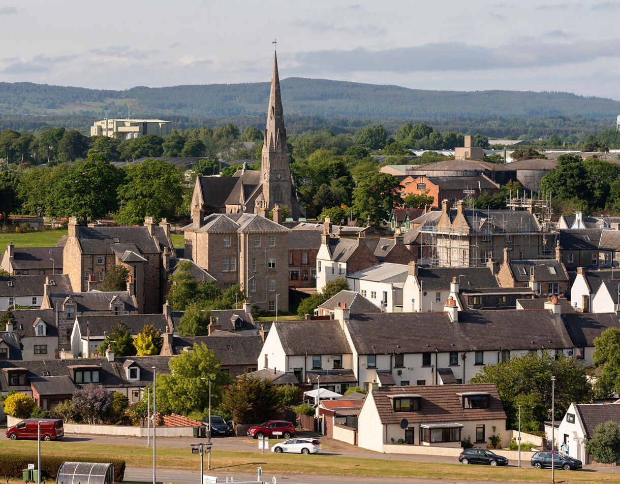 A panoramic view of Invergordon, Scotland, featuring stone houses, a church with a tall spire and wooded hills in the distance under a partly cloudy sky.