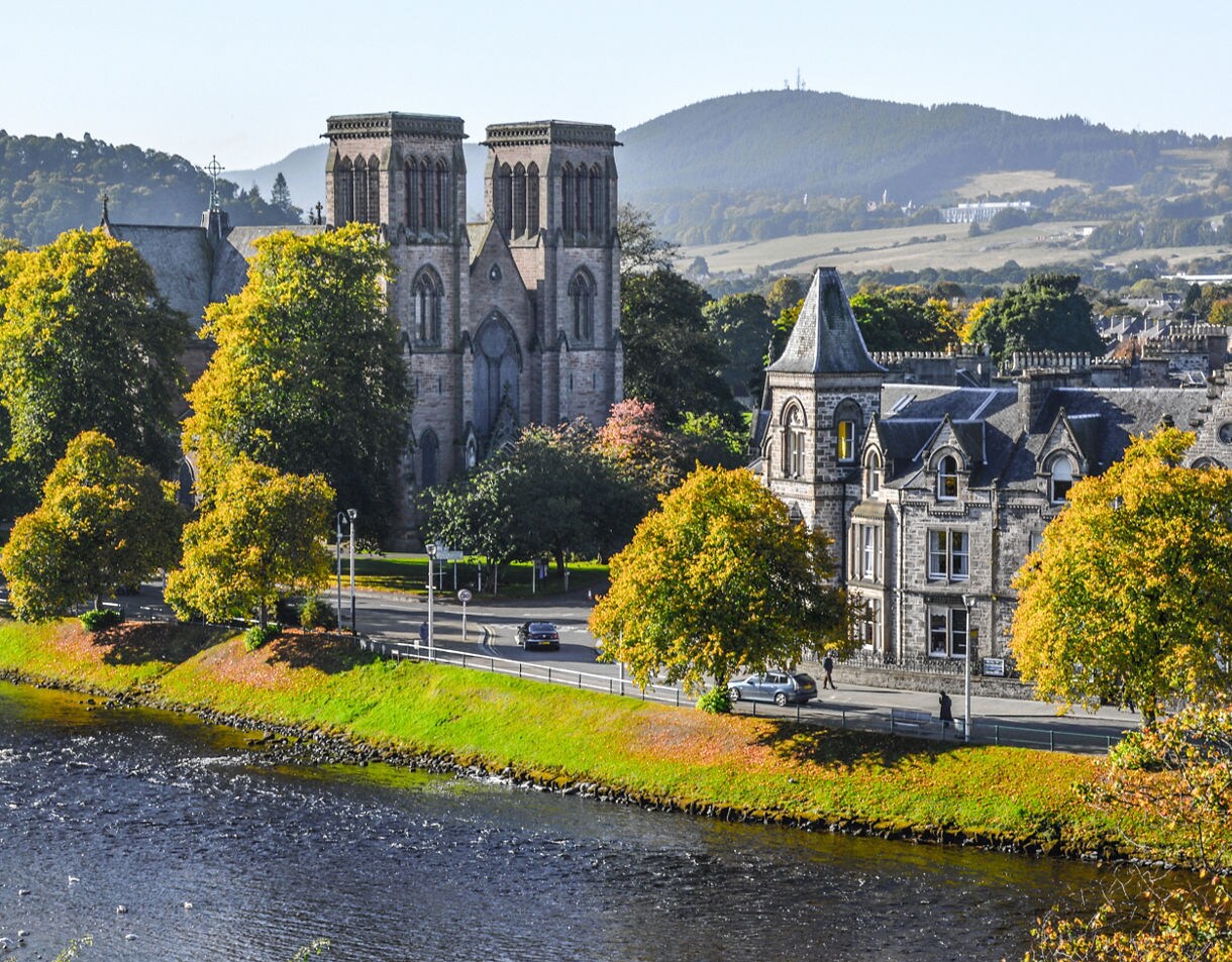 View of Inverness Cathedral and historic stone buildings along the River Ness, surrounded by trees with autumn foliage and hills in the background.