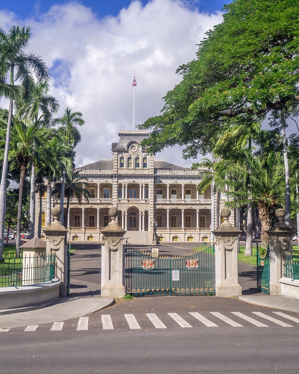 Iolani Palace in Honolulu, a historic royal residence surrounded by palm trees, ornate gates and lush green lawns under a bright blue sky.