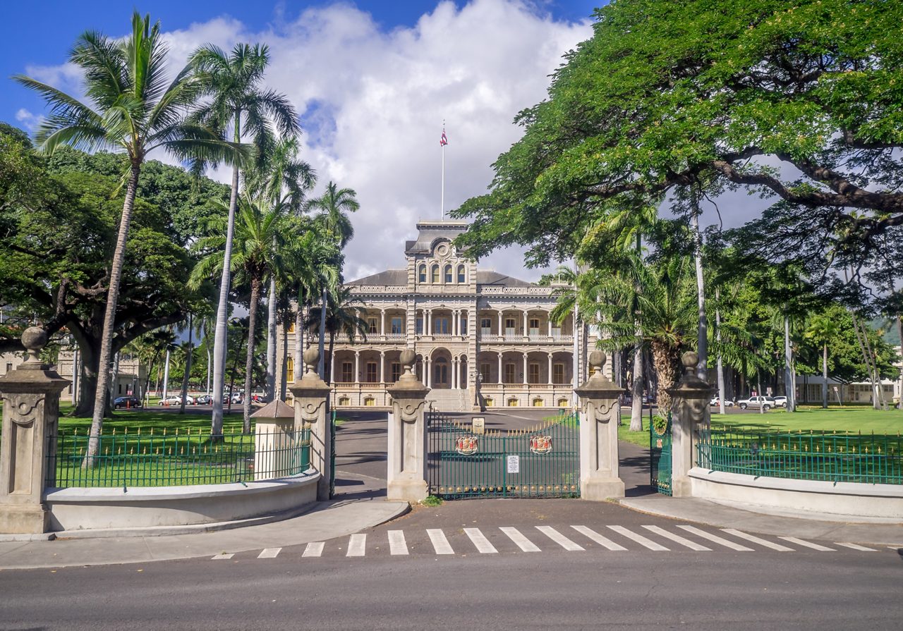 Iolani Palace in Honolulu, a historic royal residence surrounded by palm trees, ornate gates and lush green lawns under a bright blue sky.