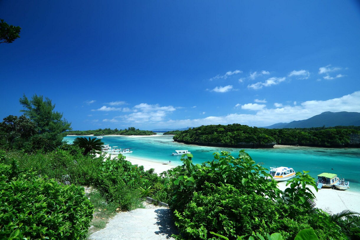 Aerial view of Kabira Bay in Ishigaki Island showing crystal clear turquoise water, white sandy beaches surrounded by lush green vegetation, several small boats anchored in the bay, and mountains visible in the background under a bright blue sky with white clouds.