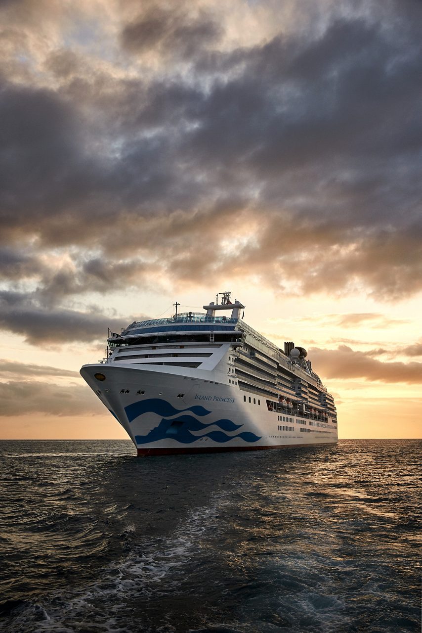 The Island Princess cruise ship sailing at sea during sunset, with a cloudy sky casting a warm glow over the water and the ship’s sleek white hull standing out against the golden horizon.