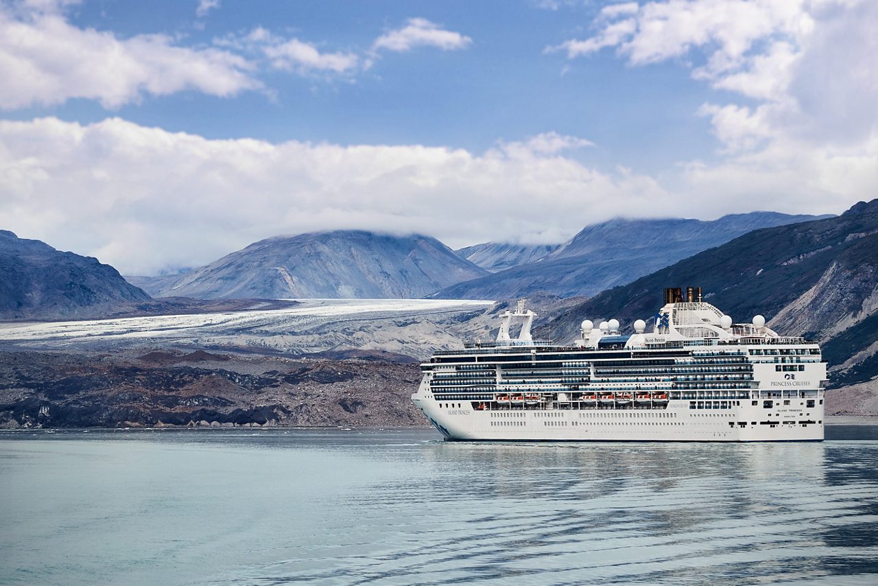 he Island Princess cruise ship sailing through the serene waters of Glacier Bay National Park in Alaska, with rugged mountains and a glacier in the background under a partly cloudy sky. 