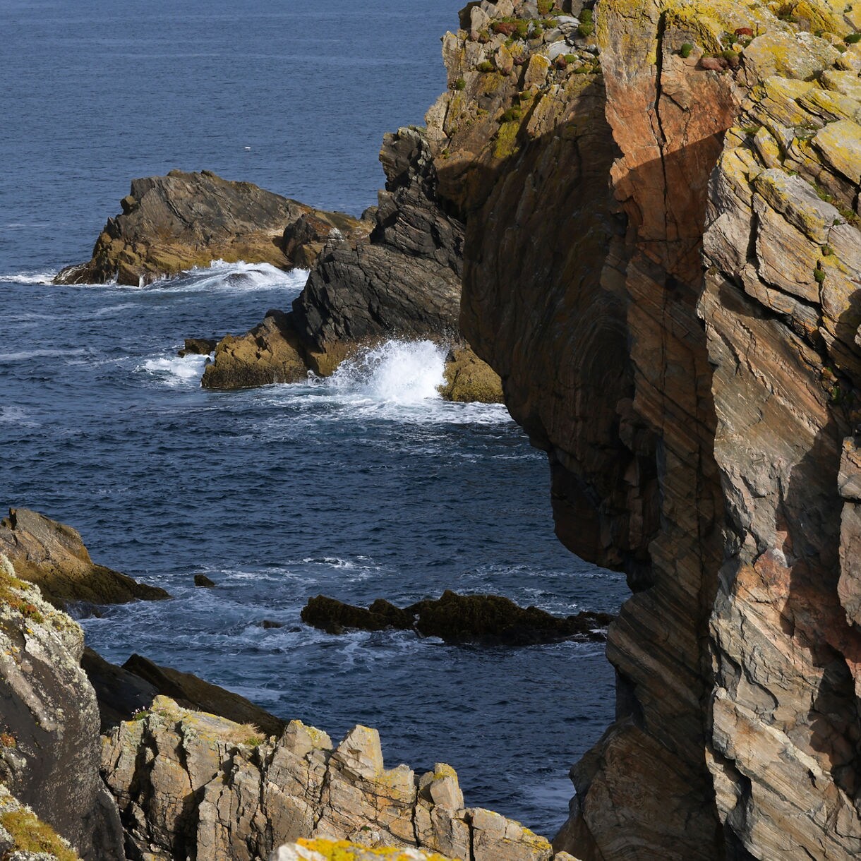 Rocky sea cliffs on the Isle of Lewis with waves breaking against jagged formations, patches of grass and lichen covering the edges and deep blue water swirling below.