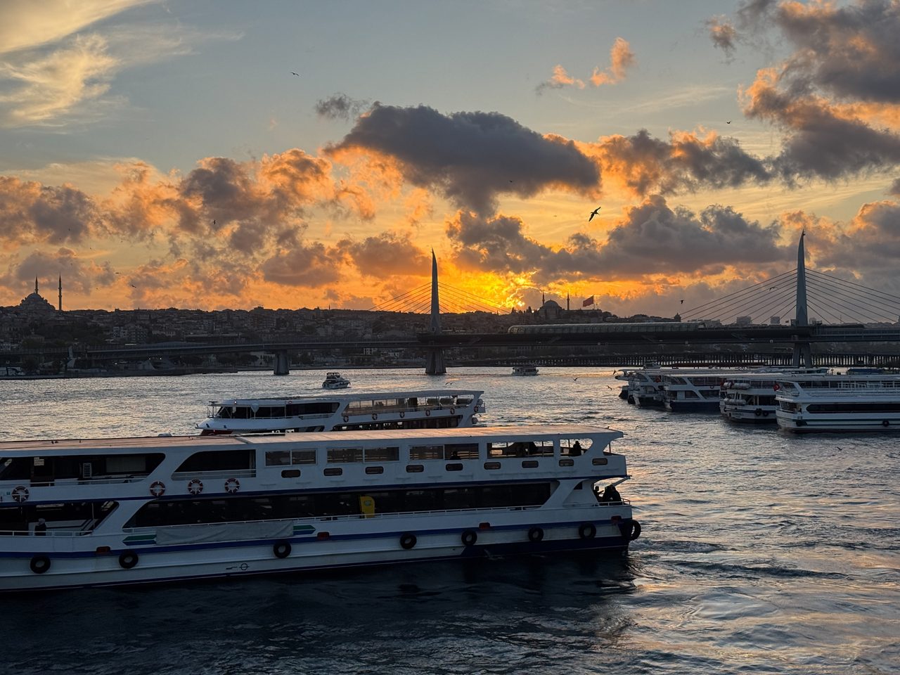 Sunset over Istanbul with ferries on the Bosphorus, a modern bridge, and mosque silhouettes against fiery clouds.