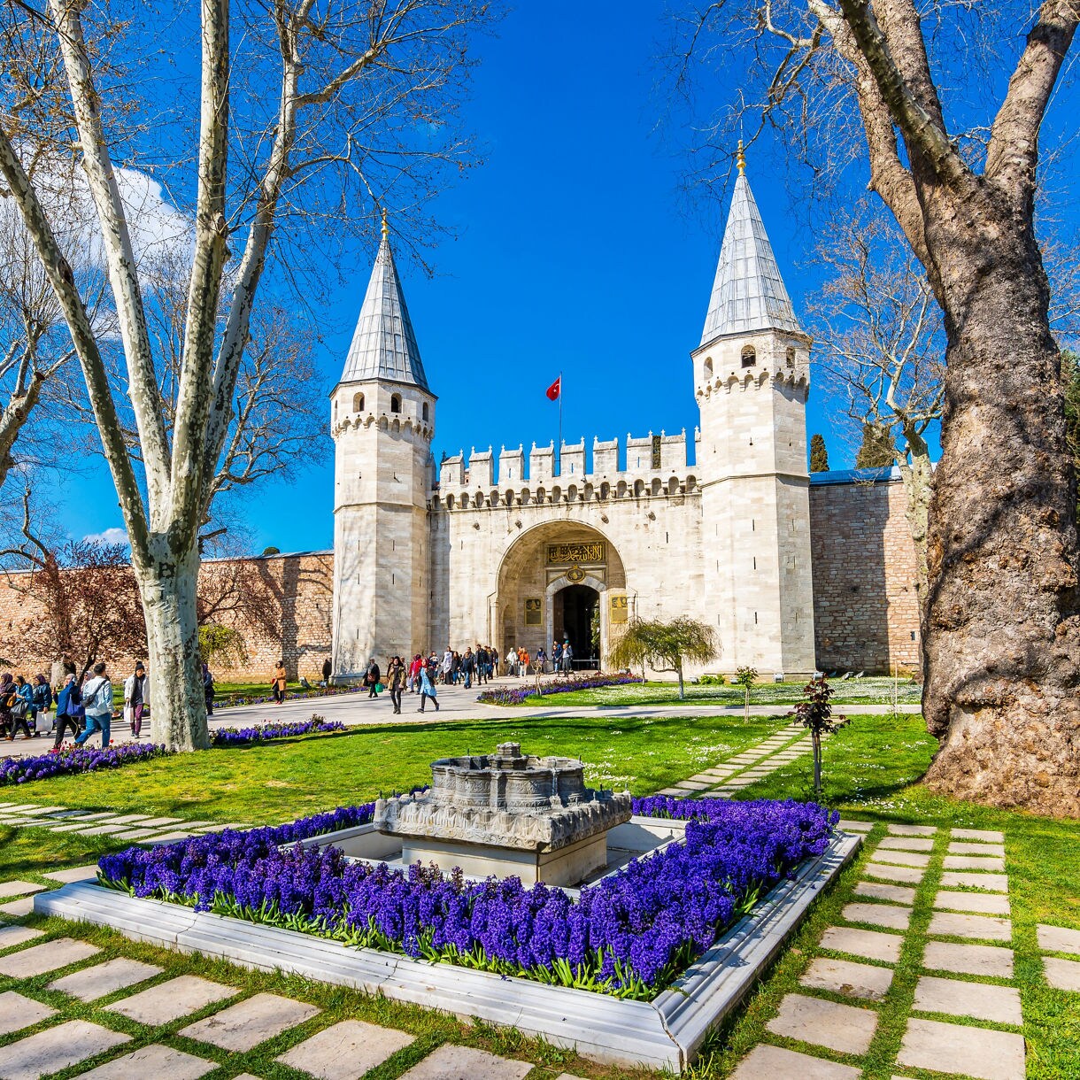 Entrance to Istanbul’s Topkapi Palace with twin towers, arched gateway, and gardens lined with purple flowers.