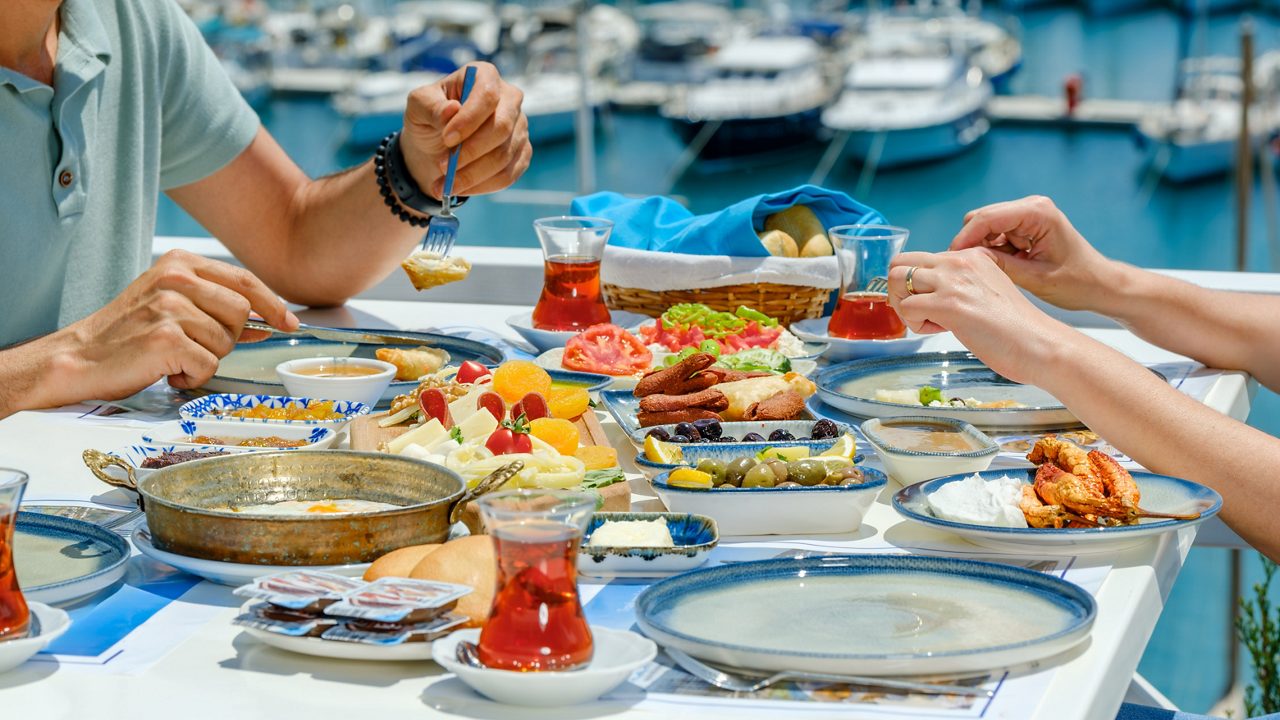 Table set with Turkish breakfast dishes including olives, cheeses, tomatoes, pastries, and glasses of tea beside a marina.