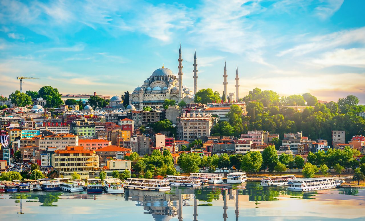 Panoramic view of Istanbul with the Suleymaniye Mosque rising above colorful hillside buildings and boats docked along the Bosphorus.