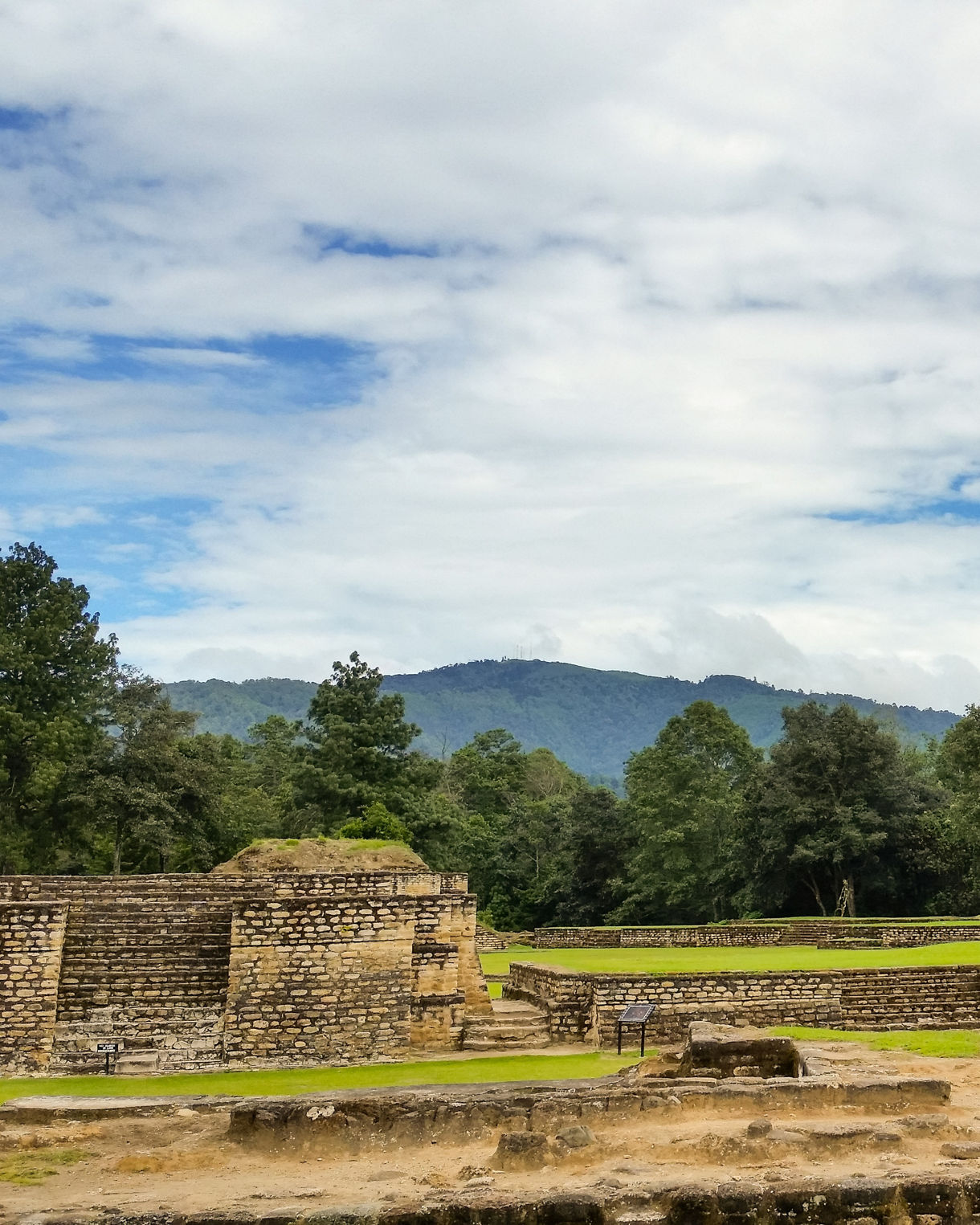 Stone structures and grassy terraces of the Iximché archaeological site in Guatemala, set against a backdrop of forested hills and a partly cloudy sky.
