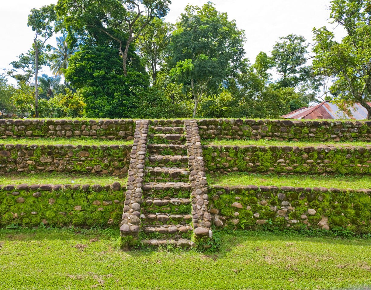Stone steps leading up a tiered, mossy pyramid structure at the Izapa archaeological site, surrounded by lush green trees.