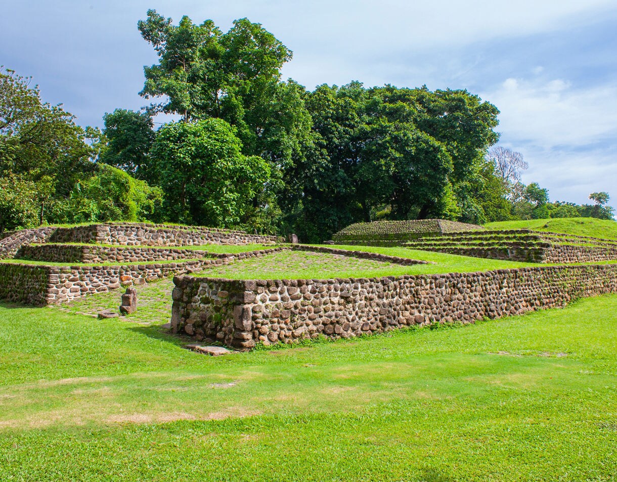 Archaeological site of Izapa in Chiapas, Mexico, showing low stone pyramids and terraces surrounded by vibrant green grass and large tropical trees.