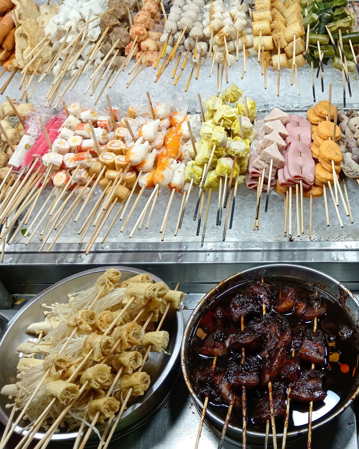 Assorted skewered meats, seafood and vegetables displayed on ice and simmering in sauces at a Malaysian street food stall.