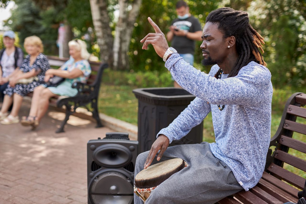 Jamaican musician with dreadlocks playing traditional drum while seated on bench with tourists watching in Falmouth town park.