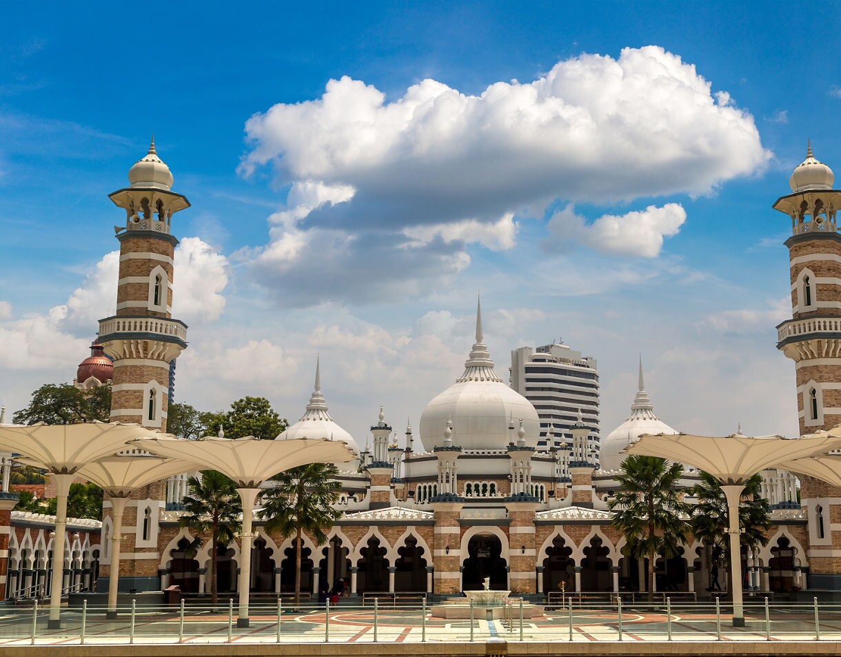 Masjid Jamek mosque in Kuala Lumpur with white domes, striped minarets and arched entrances against a blue sky.