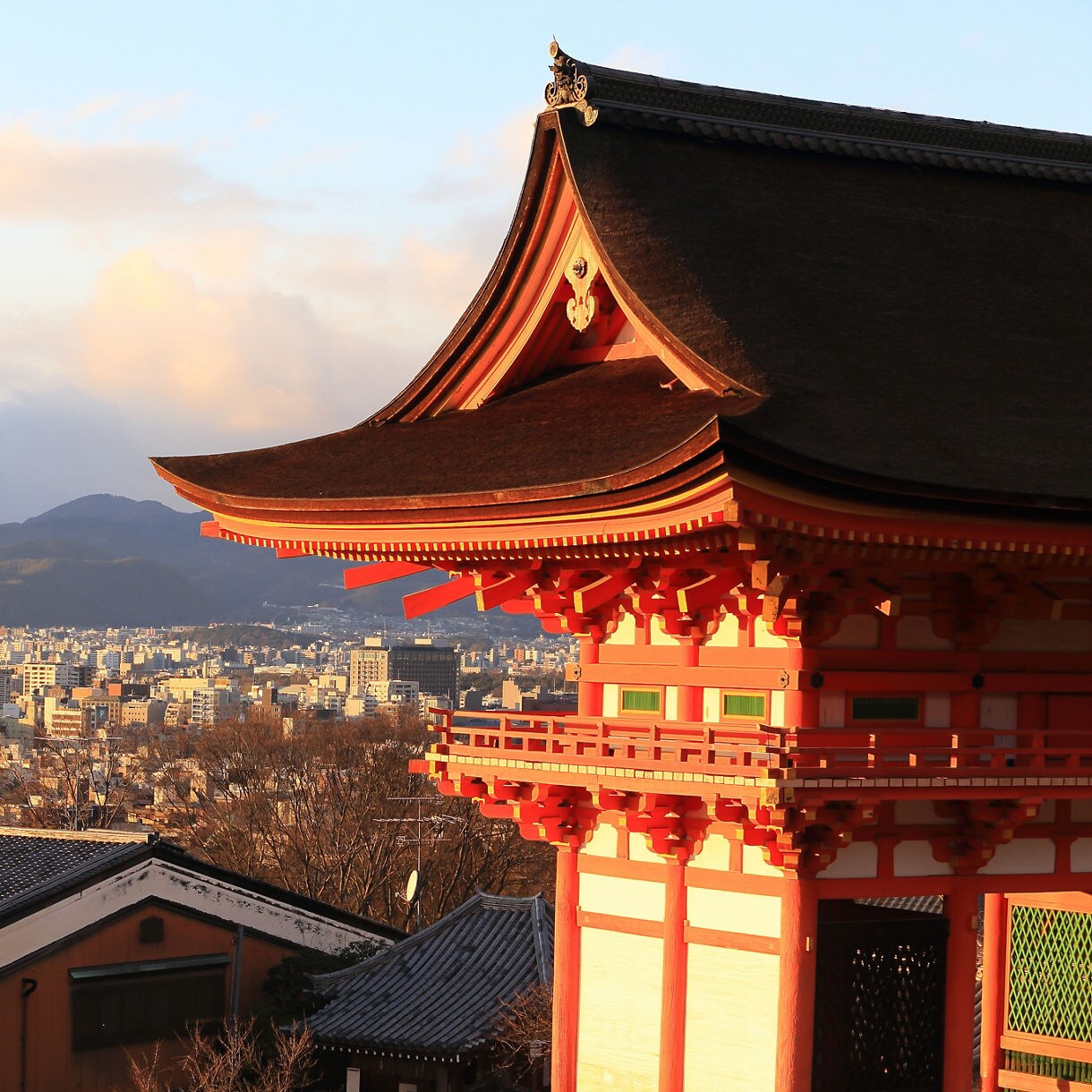 The vivid vermilion gate of Kiyomizu-dera Temple glows in warm sunset light, with a sweeping panorama of Kyoto's cityscape and rolling green mountains stretching into the distance beneath a dramatic sky.