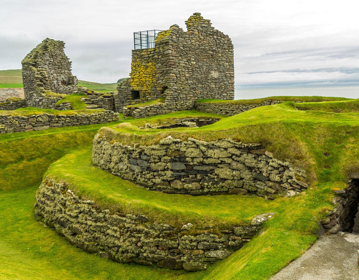 Stone ruins and circular walls of Jarlshof settlement in Shetland, surrounded by green grass with the sea in the background.