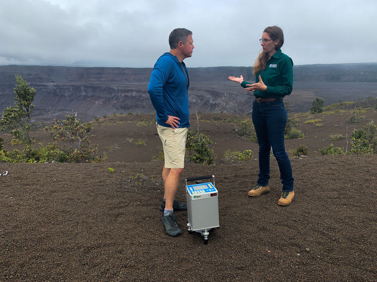Jeff Corwin talks with a researcher holding scientific equipment on a volcanic landscape.