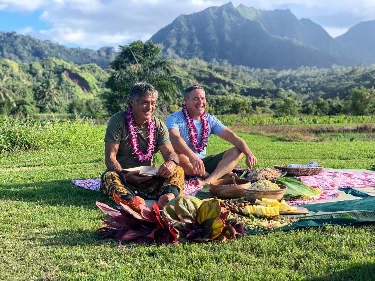 Two men wearing flower leis sit cross-legged on grass beside a spread of traditional food, with mountains behind them.