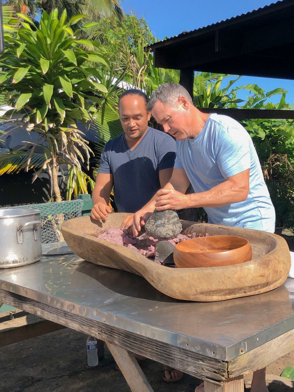 Two men use stone tools to prepare food in a large wooden bowl on an outdoor table.