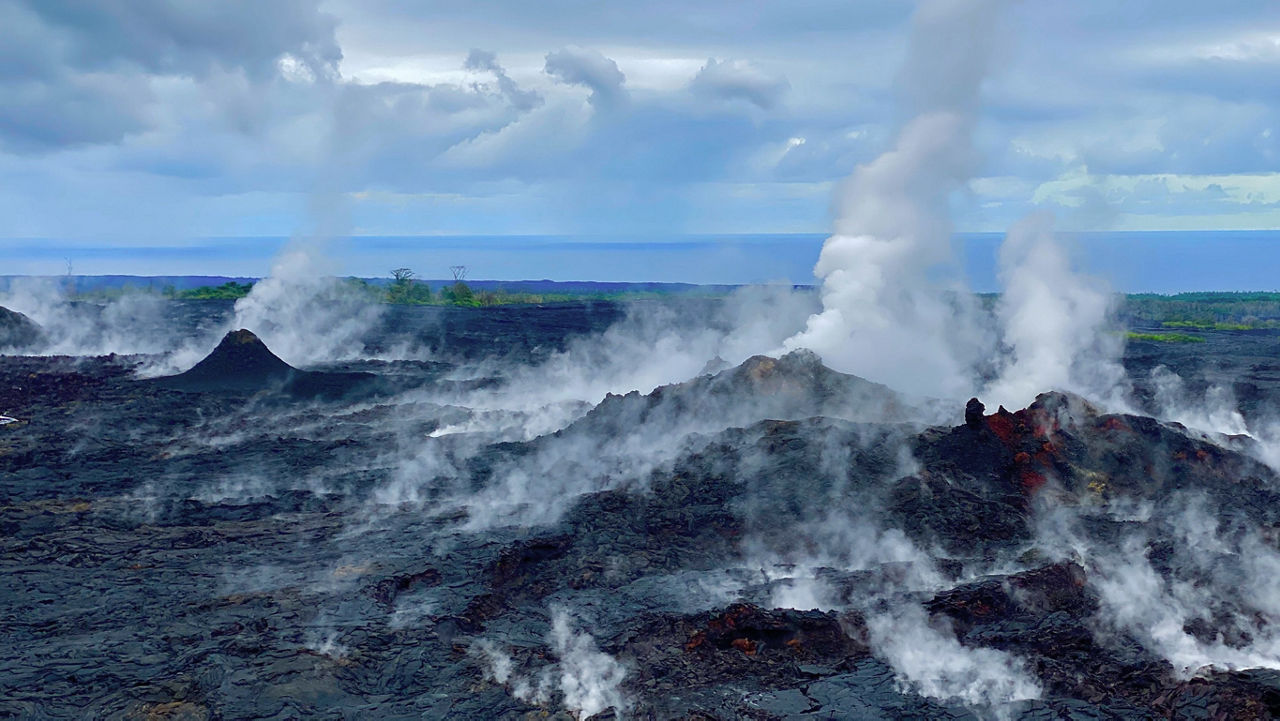 Thick steam and smoke rise from a volcanic crater with the ocean visible in the distance.