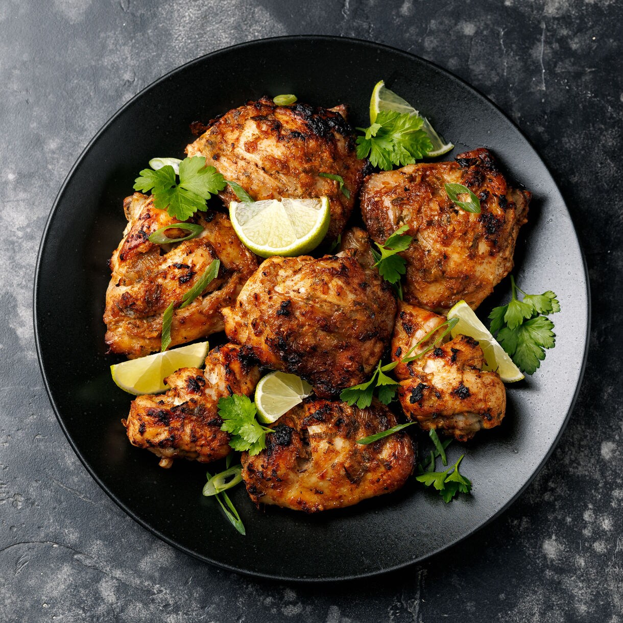 Plate of Jamaican jerk chicken garnished with lime wedges and cilantro on a dark background.