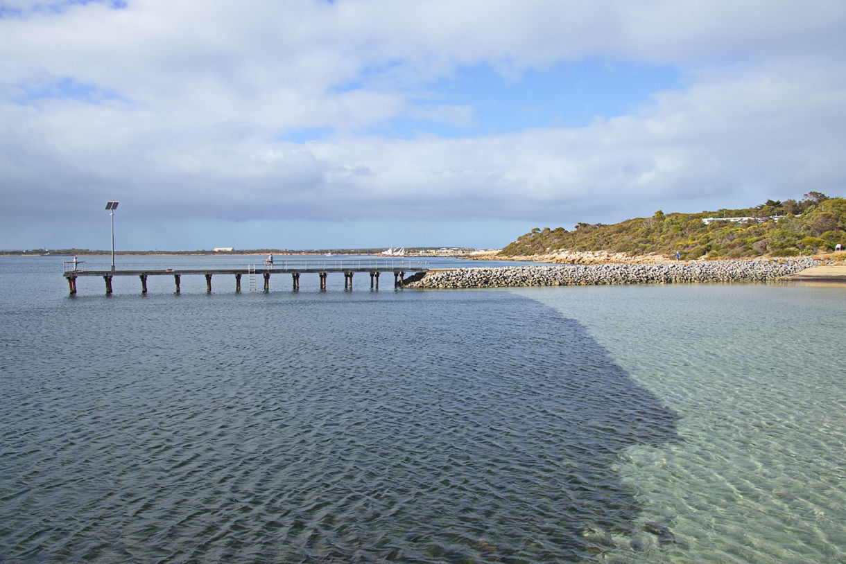 A narrow pier extends over calm, rippled water toward a rocky shoreline with low shrubs under a partly cloudy sky.