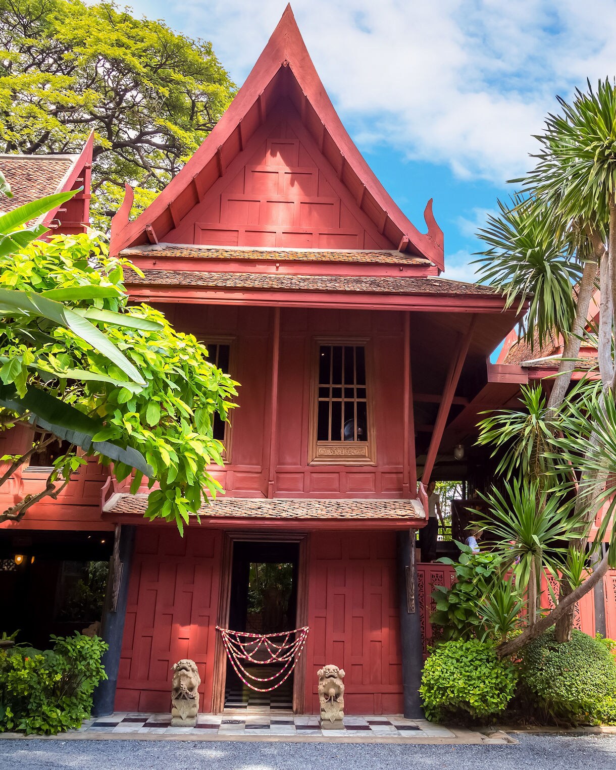 Red wooden Thai-style house with steep gabled roofs surrounded by palm trees and greenery under a bright sky.