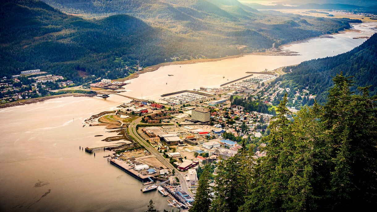 Aerial view of Juneau, Alaska, with its harbor, colorful buildings and rugged mountains towering in the background under a glowing sunset sky.
