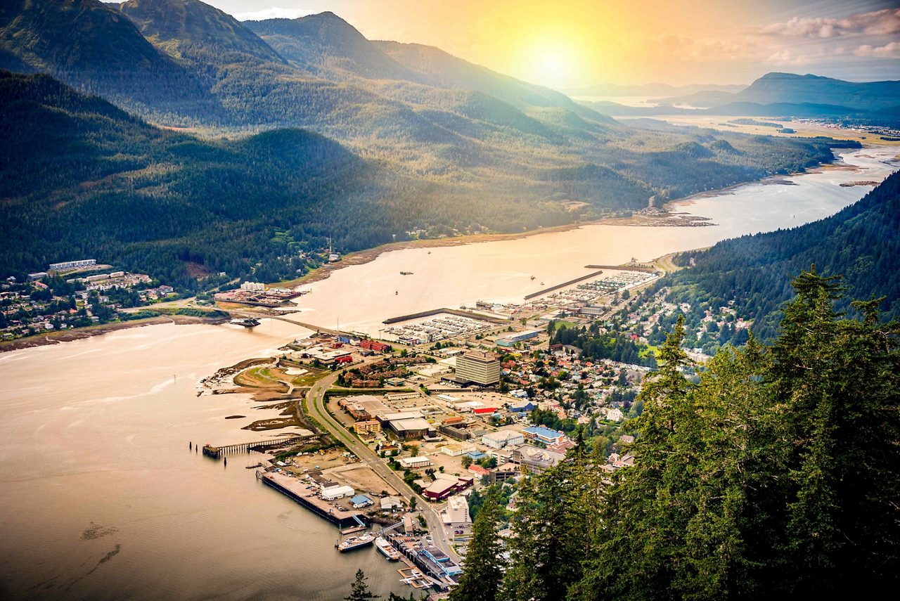 Aerial view of Juneau, Alaska, with its harbor, colorful buildings and rugged mountains towering in the background under a glowing sunset sky.