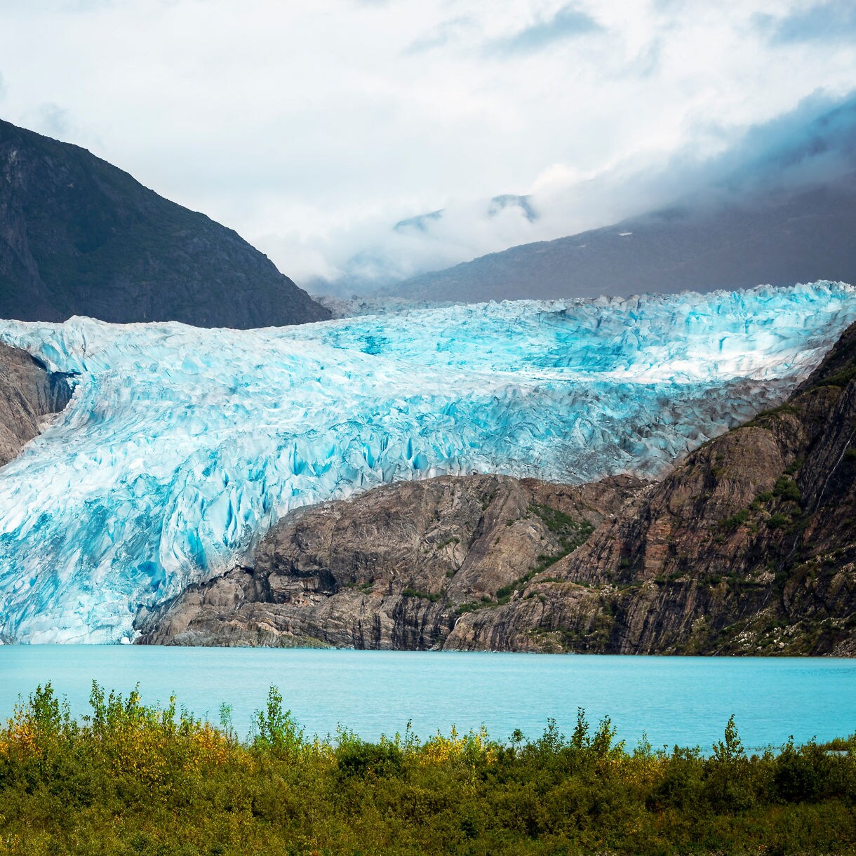 View of Mendenhall Glacier in Juneau, Alaska, with blue ice flowing down rocky cliffs into a turquoise lake surrounded by lush greenery and distant mountains.