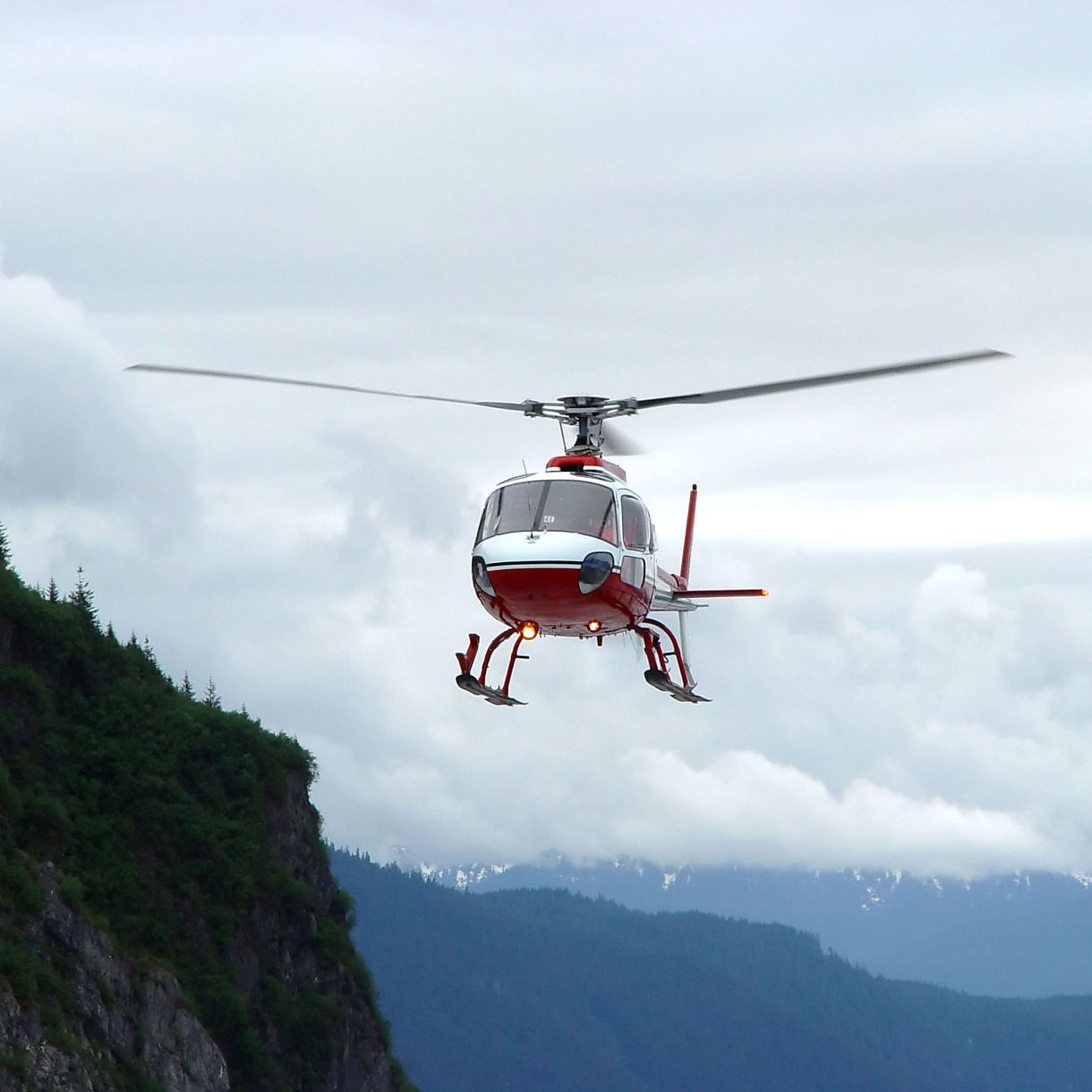 A helicopter flying over the cliffs near Mendenhall Glacier, with green forests below and snow-capped mountains in the distance under a cloudy sky.
