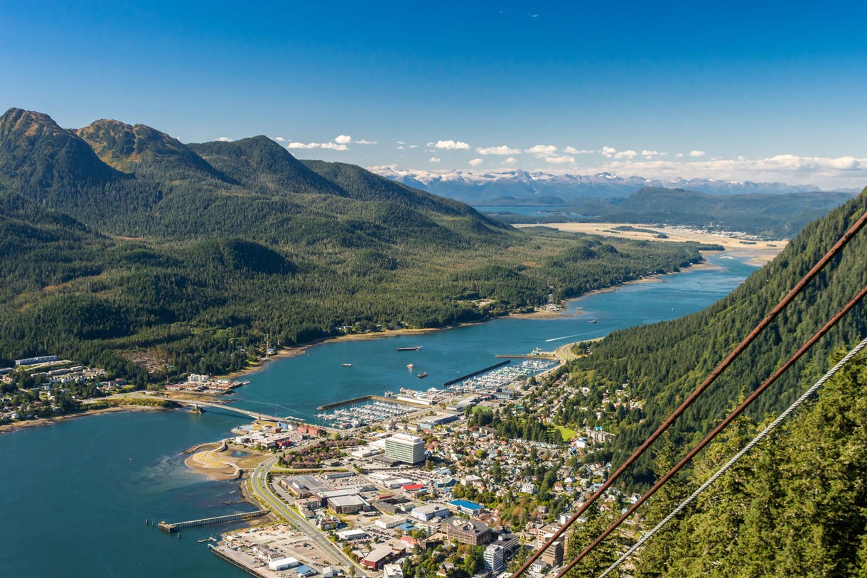 An aerial view of Juneau, Alaska, taken from Mount Roberts Tramway, showcasing the city, cruise ship port, and surrounding green mountains with clear blue skies and waterways in the distance.