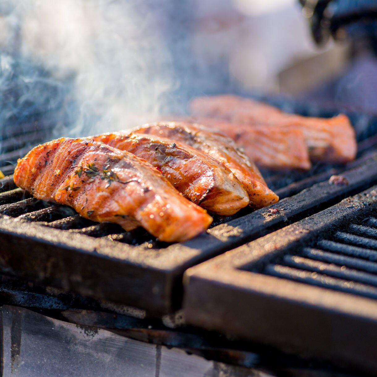 Close-up of salmon fillets grilling on a barbecue with lemon wedges and a cob of corn, surrounded by smoke, during a Juneau salmon bake.