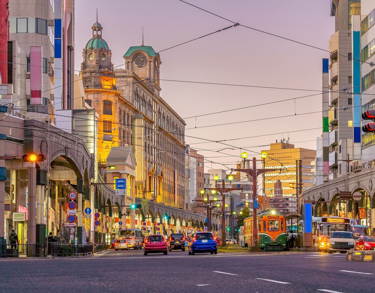 Evening view of downtown Kagoshima, Japan with cars, buses and a retro green tram traveling through a lively street lined with shops and lit buildings.