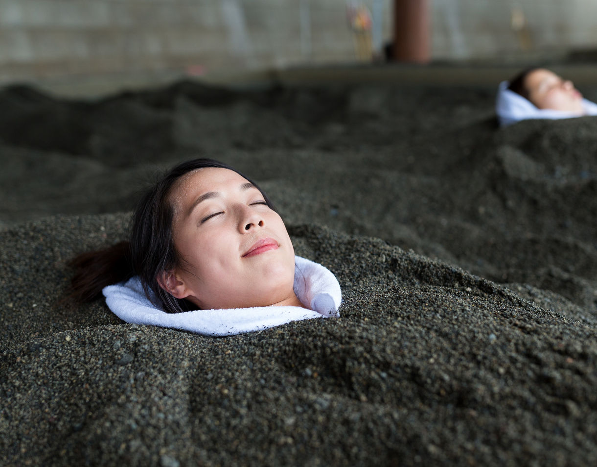 Woman buried in warm black sand up to her neck with a towel around her shoulders, relaxing during a hot sand bath in Ibusuki, Japan.