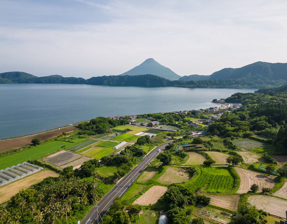 Aerial view of Lake Ikeda in Kagoshima, Japan, with green fields, scattered homes and Mt. Kaimon’s cone-shaped peak in the background.