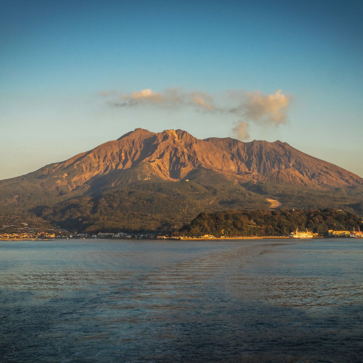 Sakurajima volcano viewed from the water, with smoke rising from its peak and the coastline of Kagoshima visible below.