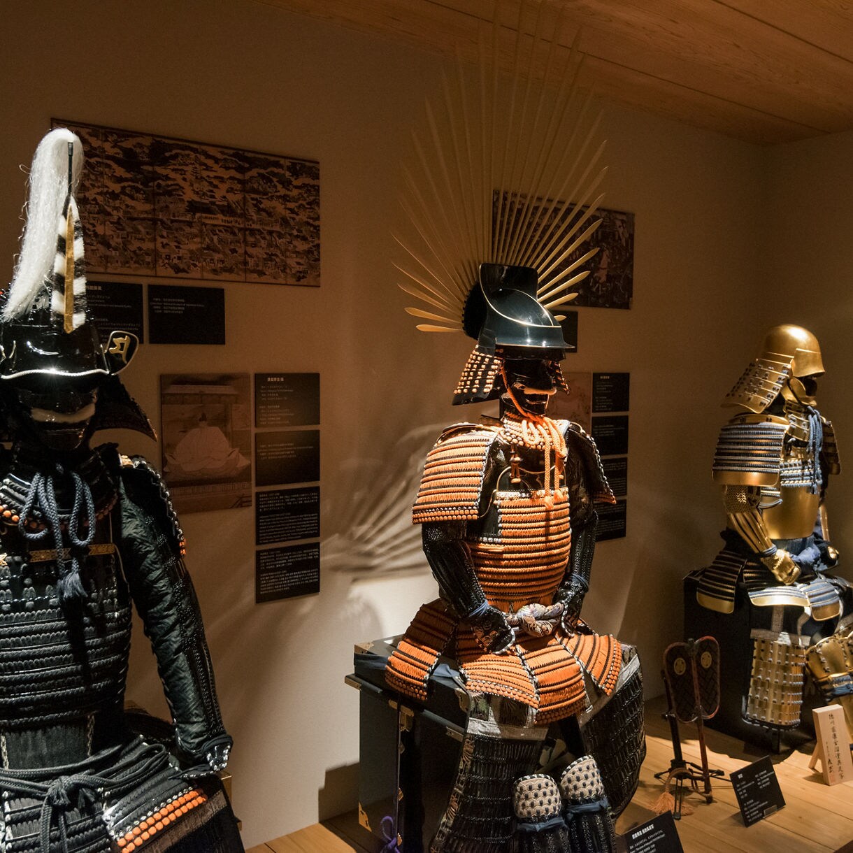 Three traditional samurai armors displayed in a museum, each with distinct helmets and ornate designs.
