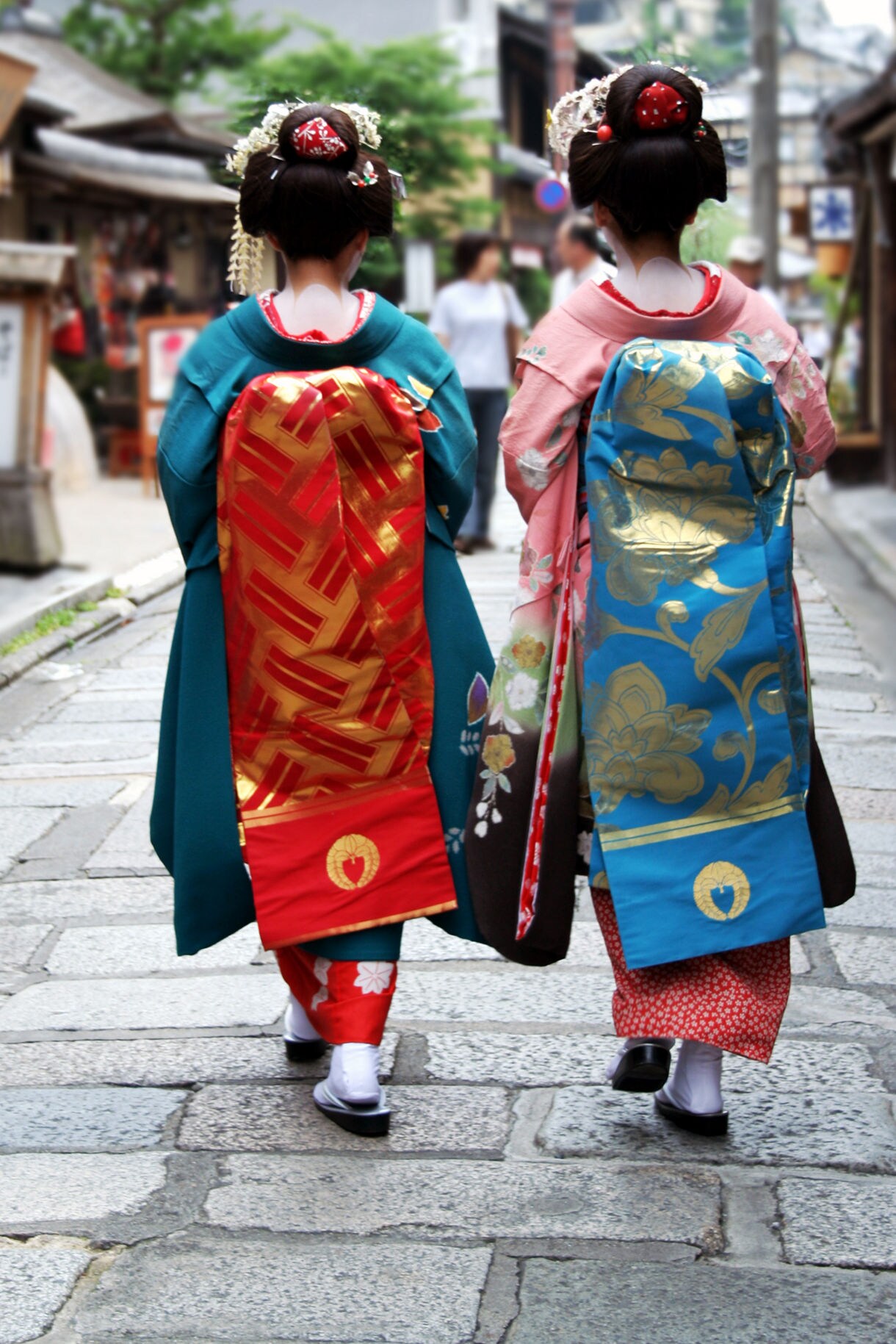 Two women wearing traditional Japanese kimono with elaborate hair ornaments walk away from the camera down a stone-paved street in Kanazawa's historic district, one in a teal kimono with red and gold obi, the other in pink with a blue and gold obi, with traditional Japanese buildings visible in the background.