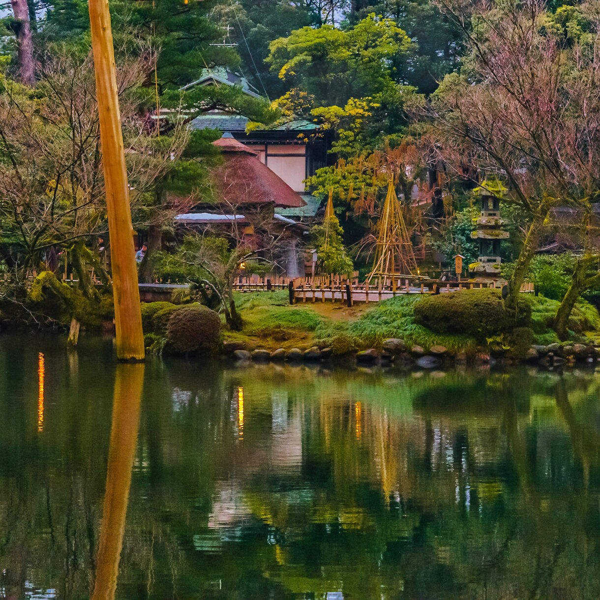 Twilight view of Kenrokuen Garden with a pond, stone lantern, bare trees and buildings softly lit among mossy greenery.