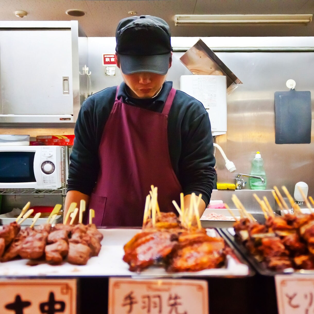 Vendor in apron and cap standing behind trays of grilled meat skewers inside a small kitchen at Omicho Market.