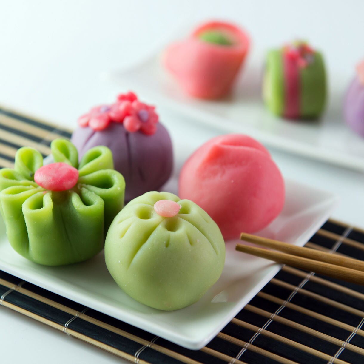 Colorful Japanese wagashi sweets shaped like flowers on white plates with chopsticks and a bamboo mat.