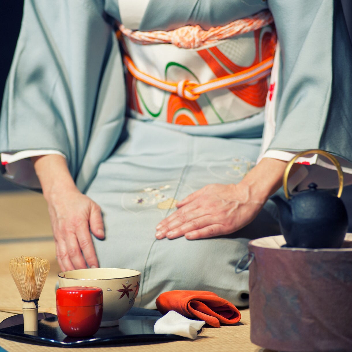 Person in a kimono kneeling on tatami mat next to tea utensils, including a whisk, bowl and iron kettle.