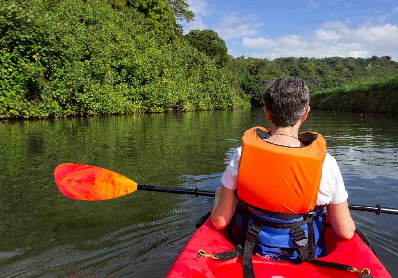 A person wearing an orange life vest paddles a red kayak along a calm, tree-lined waterway.