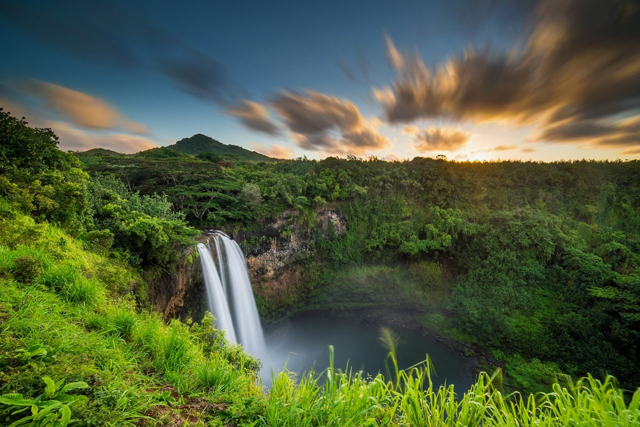 Wailua Falls in Kauai, Hawaii, captured at sunrise with water flowing into a serene pool below, surrounded by lush tropical greenery and a vibrant sky with streaks of colorful clouds. 