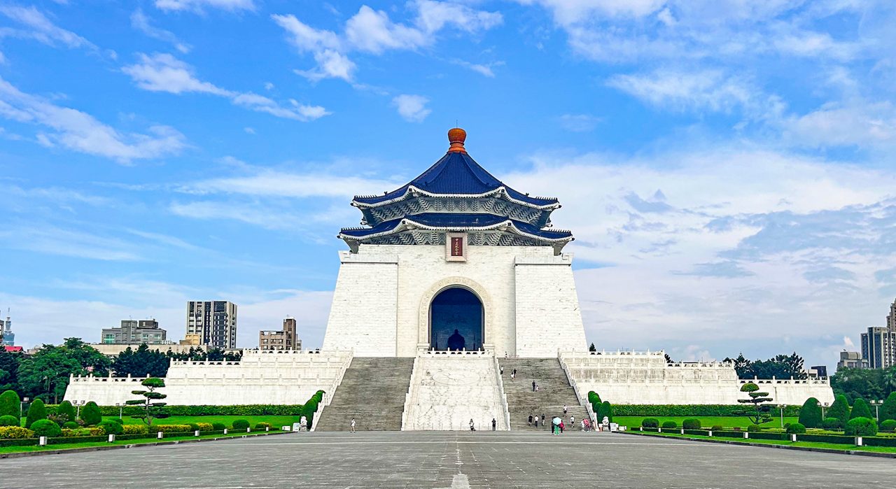 The white marble Chiang Kai-shek Memorial Hall with a blue-tiled octagonal roof, towering above a wide stone plaza and flanked by manicured gardens and modern Taipei buildings in the distance.