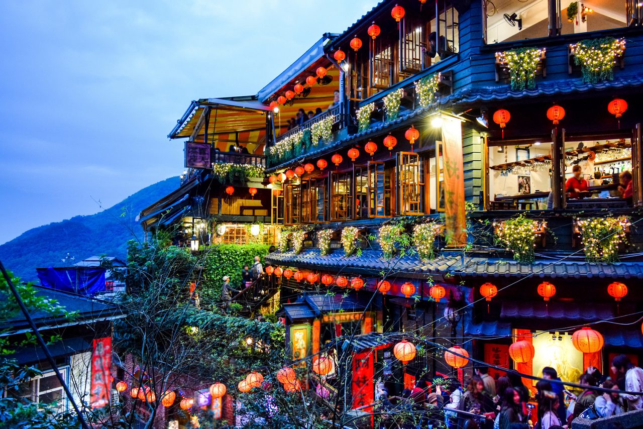 Colorful view of Chiufen Old Street in Taiwan at twilight, with glowing red lanterns, bustling crowds, traditional wooden buildings and mountainous scenery in the background.