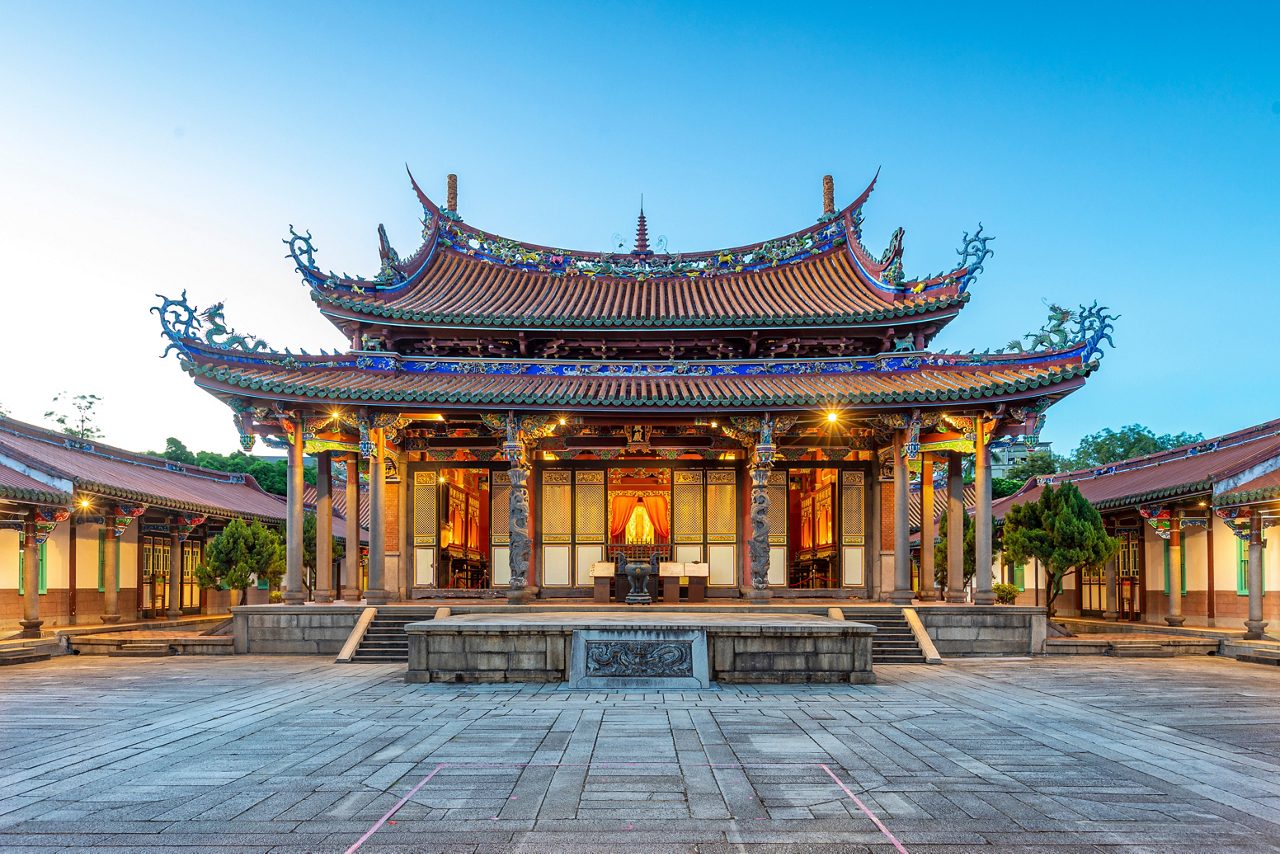 The Taipei Confucius Temple at dusk, featuring intricate carvings, colorful dragon-adorned rooftops and a grand symmetrical courtyard lined with traditional red-tiled buildings.