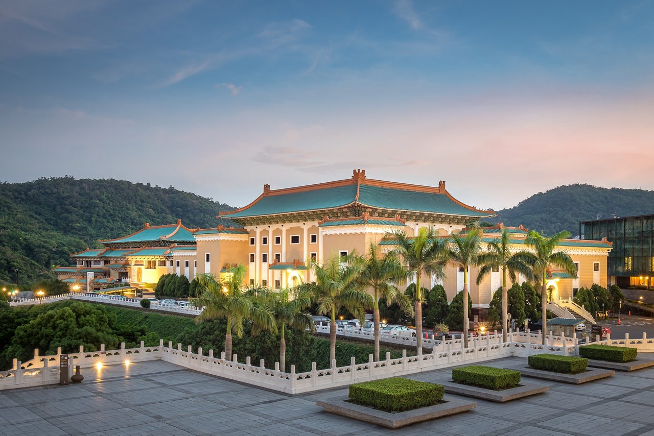 The National Palace Museum in Taipei illuminated at sunset, showcasing classic Chinese architecture with green tiled roofs, surrounded by palm trees, gardens and forested hills.