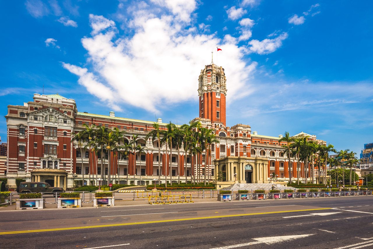 The red and white colonial-era Presidential Office Building in Taipei, Taiwan, featuring a central clock tower, rows of palm trees and a bright blue sky with scattered clouds overhead.